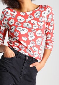 Langarm, koraalkleurig top met zwart bloemenpatroon. Heeft een brede halslijn en een getailleerd silhouet, gecombineerd met hoge zwarte jeans.