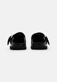 Sandales en cuir noir avec un dos ouvert, ornées d'un accent en nœud et de garnitures en métal argenté. Texture lisse et semelle plate pour un confort optimal.