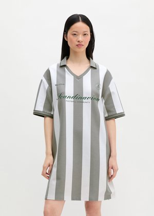 Jonge vrouw met lang zwart haar draagt een losse, grijs-wit gestreepte jurk met verticale strepen en de tekst "Scandinavian Football Community".