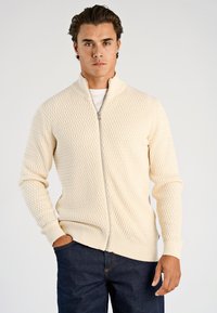 Kremowy sweter na zamek z teksturowanym wzorem diamentowym, z prążkowanymi mankietami i dołem. Połączony z ciemnymi dżinsami, stoi na jasnym tle.