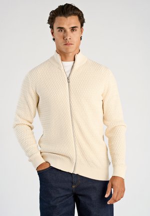 Maglione crema con cerniera e motivo a diamante testurizzato, polsini e orlo a costine. Abbinato a jeans scuri, in piedi contro uno sfondo chiaro.