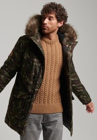Hombre con una parka de camuflaje y capucha de pelo sobre un suéter marrón de punto trenzado y jeans grises, mirando de lado frente a un fondo liso.