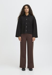 Femme avec de longs cheveux bouclés se tient vêtue d'une veste noire côtelée à boutons, d'un pantalon large marron et de chaussures noires, devant un fond clair uni.