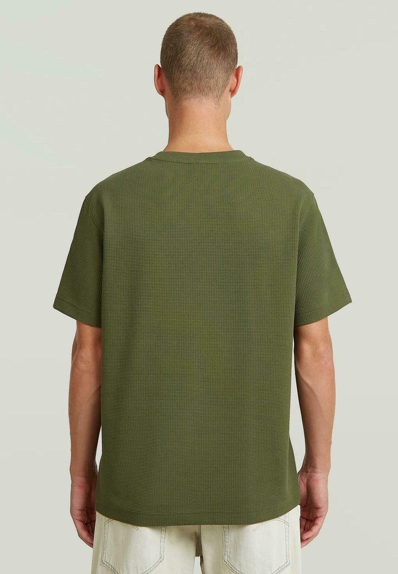 Hombre con cabello corto que lleva una camisa texturizada de manga corta color verde oliva y pantalones beige claro, de pie de espaldas sobre un fondo liso.