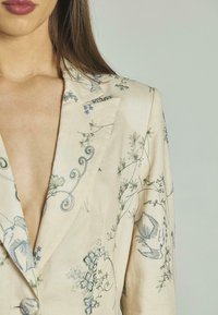 Blazer color crema con intricate ricami floreali verdi e blu, colletto a rever e silhouette aderente. Nessun bottone visibile.