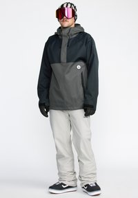 Veste de snowboard en gris foncé et noir avec un design à demi-zip, assortie à un pantalon de neige gris clair et des bottes noires. Comprend des lunettes de protection et des gants.