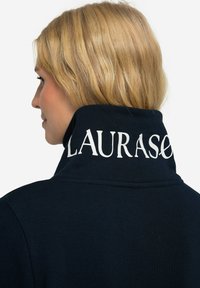 Felpa blu navy con colletto alto, caratterizzata da scritte bianche "LAURASED" sul retro. Il materiale appare morbido con una texture liscia.