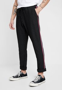 Zwarte broek met krijtstreep en een taille met trekkoord, met rode en blauwe strepen aan de zijkant. Opgerolde pijpen en gedragen met zwarte sneakers.