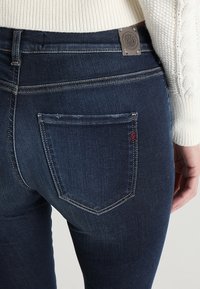 Vaqueros de mezclilla de un azul oscuro con una textura suave, que presentan un pequeño detalle de logotipo rojo en el bolsillo trasero y una etiqueta de tela en la cintura.