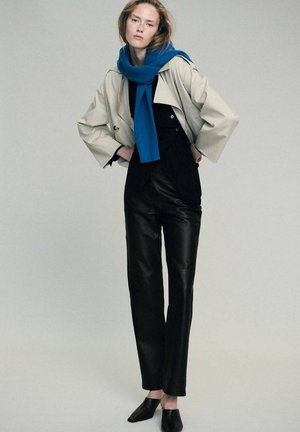 Femme debout, les mains sur les hanches, portant une veste beige, une écharpe bleue, un haut noir, un pantalon en cuir noir et des chaussures noires sur fond gris.