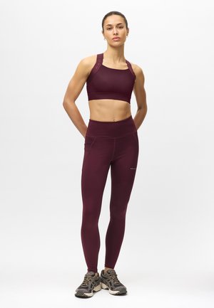 Vrouw staand in bordeaux sportbeha en leggings met grijze hardloopschoenen tegen een effen witte achtergrond.