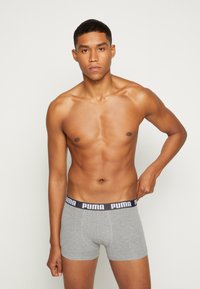 Graue Baumwollboxershorts mit einem schwarzen Puma-Logo-Gummibund. Figurbetontes Design, weiche Textur und mittellange Beine. Das Modell posiert mit einer Hand auf dem Gummibund.