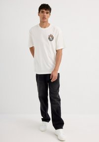 Camiseta blanca de manga corta con un gráfico de llamas coloridas en el lado izquierdo del pecho, combinada con unos jeans oscuros y zapatillas blancas.