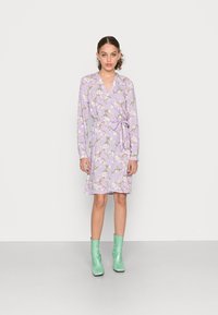 Vestido envolvente lavanda de mangas curtas com padrão floral, cinto na cintura e decote em V. Estilizado com botas verdes de tornozelo e colocado em frente a um fundo liso.