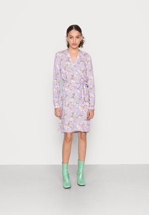 Vestido envolvente lavanda de mangas curtas com padrão floral, cinto na cintura e decote em V. Estilizado com botas verdes de tornozelo e colocado em frente a um fundo liso.