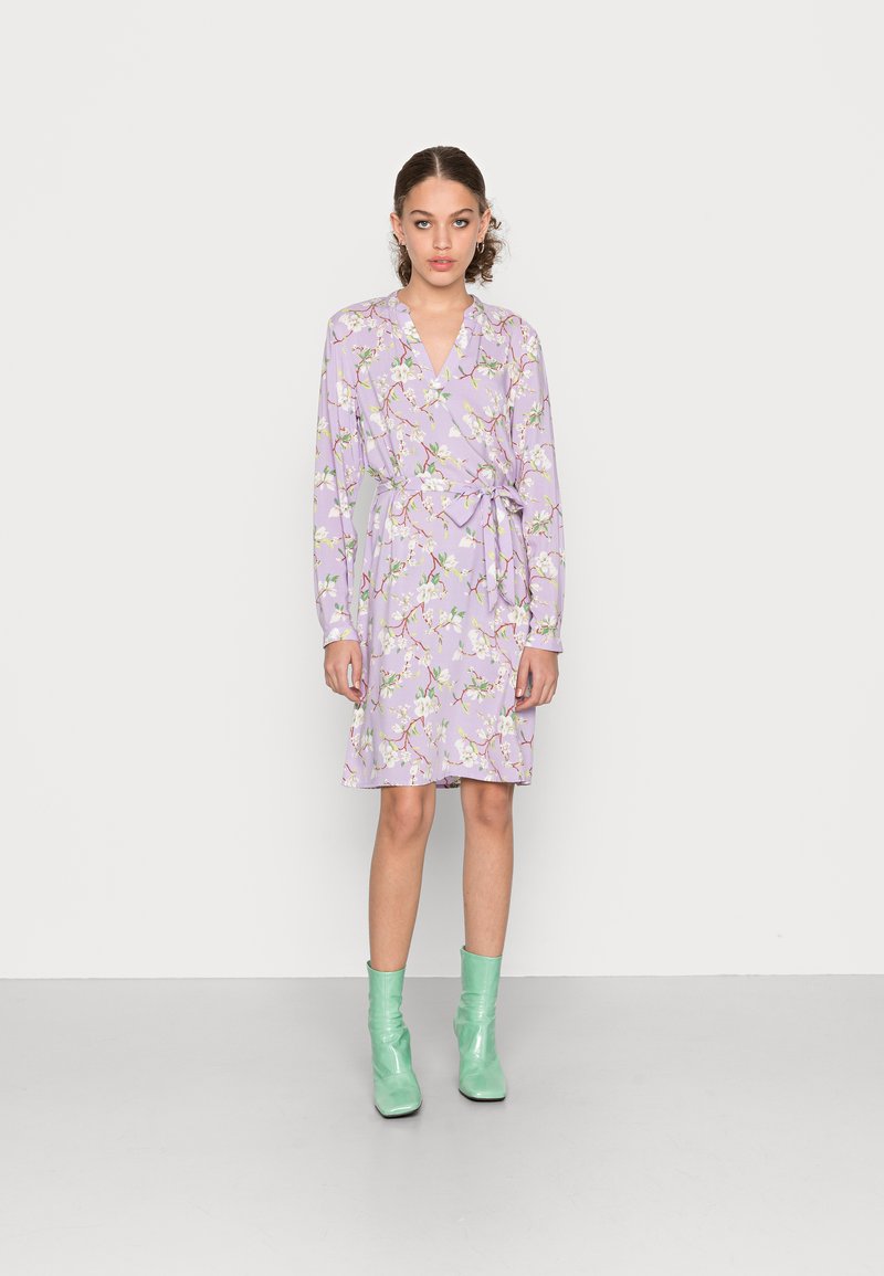 Vestido envolvente lavanda de mangas curtas com padrão floral, cinto na cintura e decote em V. Estilizado com botas verdes de tornozelo e colocado em frente a um fundo liso.