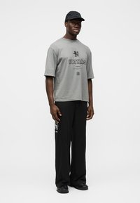 T-shirt gris à manches courtes avec le texte « ULTRAS » et un graphisme de lion. Pantalon noir avec un détail de logo. Le mannequin porte une casquette noire et des chaussures.