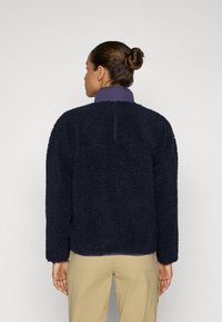 Marineblå fleecejakke med en teksturert, myk overflate, med en lilla krage og en avslappet passform. Lange ermer og avrundet søm.