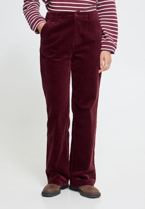 Personne portant un pantalon en velours côtelé rouge foncé avec des poches avant, associé à des chaussures marron et un haut à manches longues rayé bordeaux et blanc.