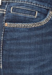 Los jeans de mezclilla azul oscuro presentan un pequeño bolsillo en la parte trasera, con un llamativo detalle de costura beige y una superficie suave y texturizada.
