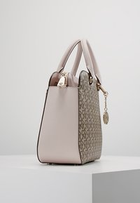 Sac à main beige à motifs avec double poignée, fermeture éclair dorée et pendentif doré suspendu, présenté sur un socle blanc contre un fond gris.
