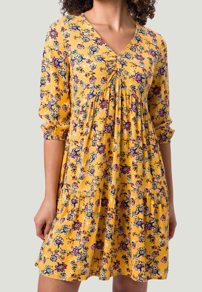 Femme portant une robe jaune à fleurs avec un col en V froncé et des manches trois-quarts sur fond gris clair.