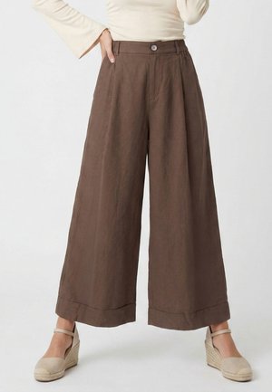 Frau trägt hoch taillierte, weit geschnittene braune Hose und beige Keil-Espadrilles, steht mit einer Hand in der Tasche vor einem schlichten Hintergrund.