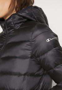 Kvinna klädd i en svart puffjacka med huva, dragkedjan stängd, med Champion-logotypen på överarmen.