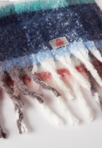 Écharpe en tricot avec un mélange de rayures bleu foncé, bleu clair et blanc, avec des franges grises et rouges. Comprend une étiquette carrée avec un logo rouge.