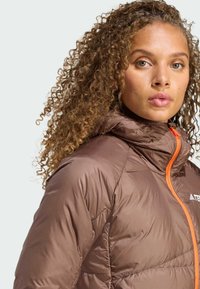 Smeđi pufasti jakna s kapuljačom, s narančastim patentnim zatvaračem i quiltanom teksturom. Istaknuti detalji uključuju logo na lijevoj strani grudi.