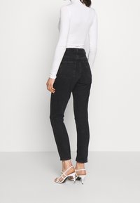 Mujer vistiendo jeans negros de tiro alto, blusa blanca de manga larga y sandalias de tacón blanco con tiras, de pie contra un fondo claro y liso.