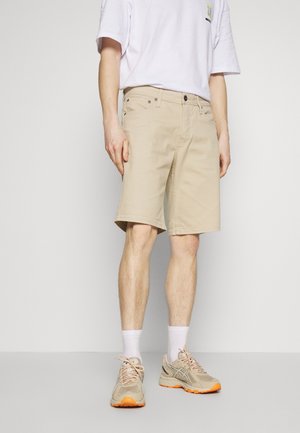 Beige puuvillashortsit, suora leikkaus ja etutaskut, yhdistettynä beigeihin lenkkareihin, joissa on oranssit yksityiskohdat ja valkoiset urheilusukat.