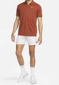 Polo Nike à manches courtes de couleur rouille, associé à un short blanc. Présente un col, un logo sur la poitrine et un tissu léger.