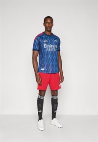 Blå fodboldtrøje med zigzag-mønster, røde detaljer, hvid "Emirates"-logo, kombineret med røde shorts og sorte sokker med hvide striber.
