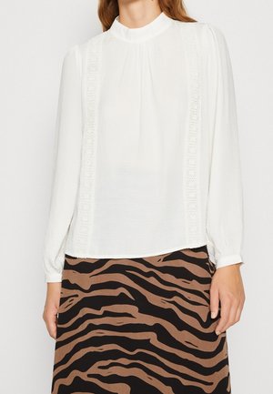 La femme porte un chemisier blanc à manches longues avec des détails en dentelle et un col montant, associé à une jupe imprimée zèbre marron et noire.