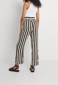 Vrouw in zwart en beige verticale gestreepte wijde broek, witte mouwloze top en zwarte sandalen, staande tegen een witte achtergrond.