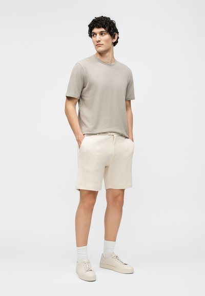 Άνδρας με μπεζ μπλουζάκι, ανοιχτό κρεμ σορτς, λευκές κάλτσες και κρεμ αθλητικά παπούτσια, στέκεται με τα χέρια στις τσέπες κοιτάζοντας αριστερά.