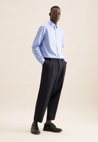 Lichtblauw gestreept overhemd met knopen, gecombineerd met donkerblauwe geplooide broek en zwarte geklede schoenen, gesitueerd tegen een neutrale achtergrond.