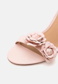 Jasnoróżowe sandały na otwartym palcu z dwoma dekoracyjnymi skórzanymi różami na pasku nad palcami.