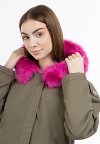 Cappotto verde oliva con un vivace cappuccio in pelliccia sintetica rosa, dotato di chiusura frontale e dettagli ai polsini. Tessuto morbido e vestibilità comoda.