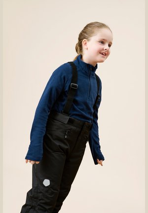 Nuori tyttö hymyilee, päällä sininen fleece-pulloveri ja mustat lumihousut, joissa on henkselit, seisten tasaisella beigen taustalla.