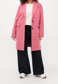 Abrigo de mezcla de lana rosa con solapas de muesca, dos bolsillos frontales y cierre de un solo botón, combinado con pantalones de pierna ancha negros y zapatillas blancas.