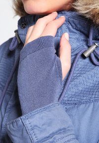 Blå jacka med quiltad textur och foder i päls, med en tumhål i ärmen för fingerlös täckning.