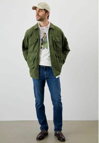 Giacca utilitaria verde con molte tasche sopra una t-shirt grafica bianca, jeans azzurri chiari e scarpe in pelle marrone. Cappello beige in testa.