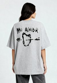 T-shirt gris surdimensionné avec un design graphique noir au dos, portant le texte "Mi Amor" et un cœur transpercé par une flèche.