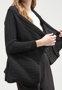 Svart stickad kofta med öppen front, texturerat mönster, långa ärmar och en lös, flödande form.