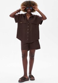 Kobieta z kręconymi włosami, ubrana w luźną brązową koszulę zapinaną na guziki, dopasowane szorty, masywne sandały i trzymająca niebieskie okulary przeciwsłoneczne nad oczami.