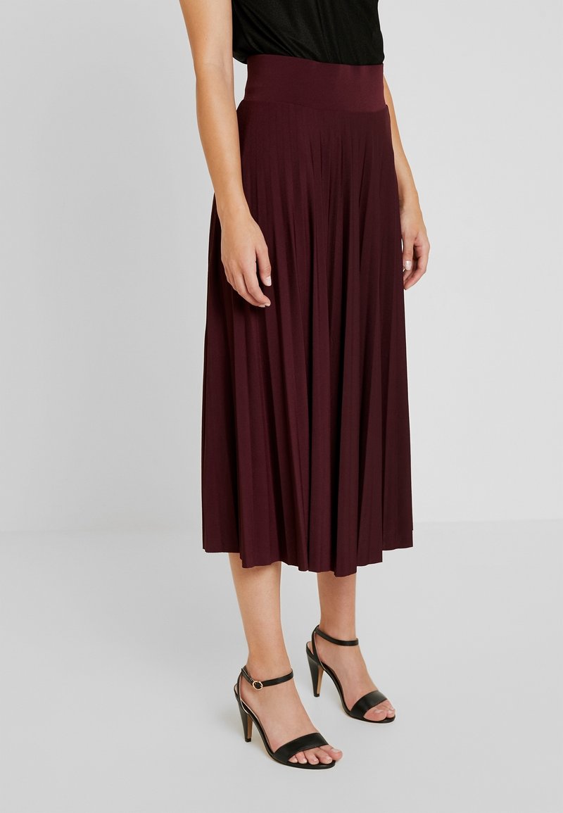 Burgundy pleated midi skirt with a wide waistband, made of soft fabric. Worn with black strappy heels, showcasing a smooth texture and drape.