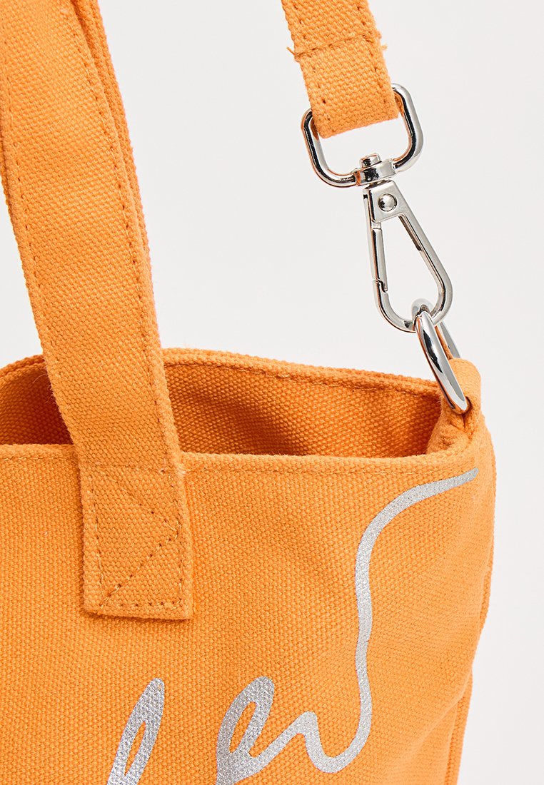 Bolso de lona naranja con asa cosida y cierre de metal plateado que conecta una correa ajustable, con texto gráfico parcial en plateado.