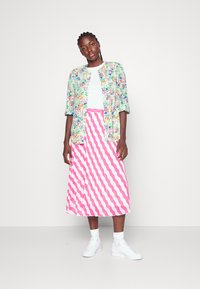 Camisa floral colorida sobre uma t-shirt branca, combinada com uma saia midi às riscas rosa e branca e ténis brancos. Texturas de tecido suaves.